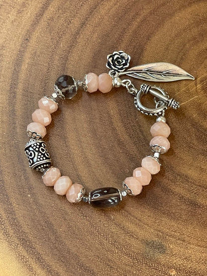 Peach moonstone and smoky quartz beaded bracelet with an ornate silver-plated focal bead, flower and leaf charms, displayed on a wooden surface.
