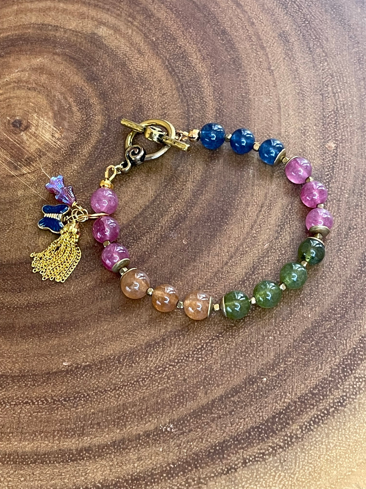 Multicolor natural tourmaline beaded bracelet with an enamel butterfly charm, gold chain tassel, and Czech flower accents, displayed on a wooden surface.