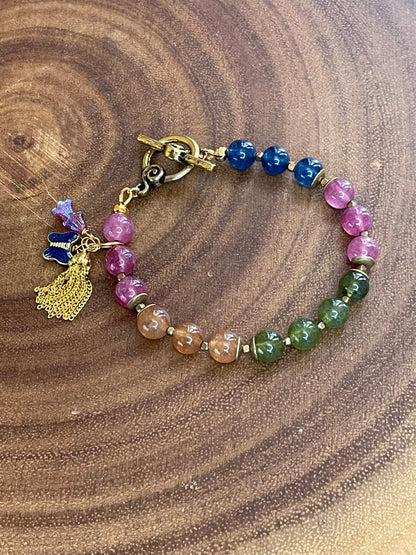 Multicolor natural tourmaline beaded bracelet with an enamel butterfly charm, gold chain tassel, and Czech flower accents, displayed on a wooden surface.