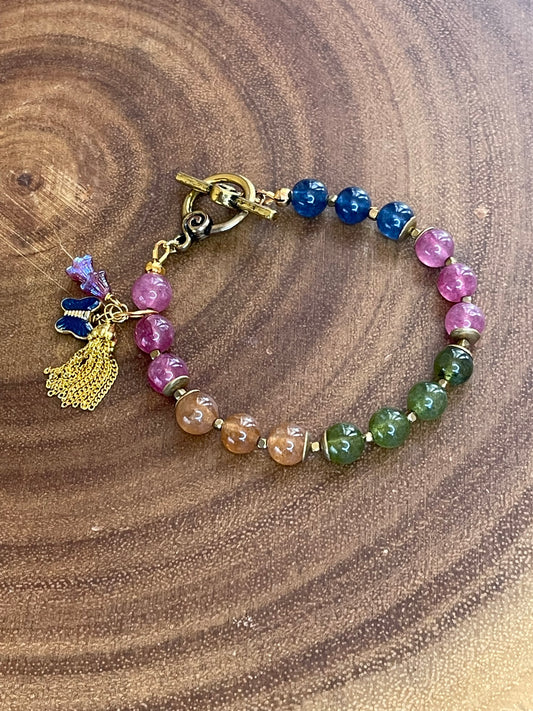 Multicolor natural tourmaline beaded bracelet with an enamel butterfly charm, gold chain tassel, and Czech flower accents, displayed on a wooden surface.