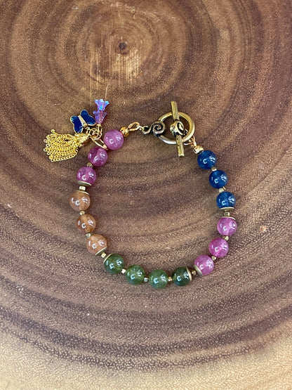 Multicolor natural tourmaline beaded bracelet with an enamel butterfly charm, gold chain tassel, and Czech flower accents, displayed on a wooden surface.