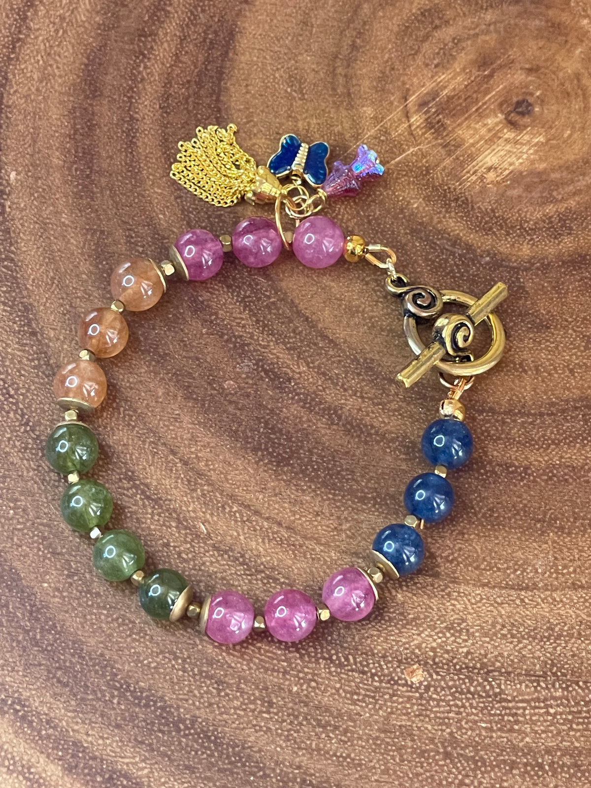 Multicolor natural tourmaline beaded bracelet with an enamel butterfly charm, gold chain tassel, and Czech flower accents, displayed on a wooden surface.