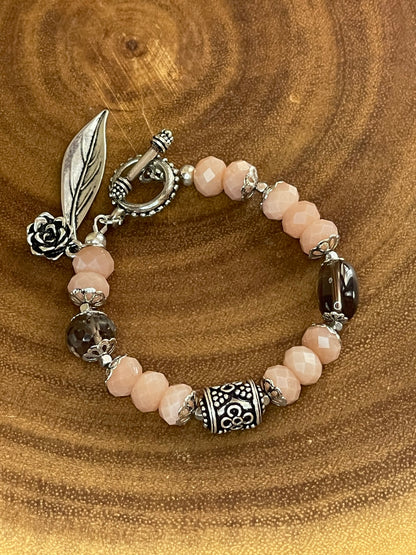 Peach moonstone and smoky quartz beaded bracelet with an ornate silver-plated focal bead, flower and leaf charms, displayed on a wooden surface.