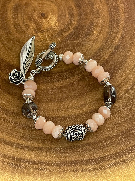 Peach moonstone and smoky quartz beaded bracelet with an ornate silver-plated focal bead, flower and leaf charms, displayed on a wooden surface.