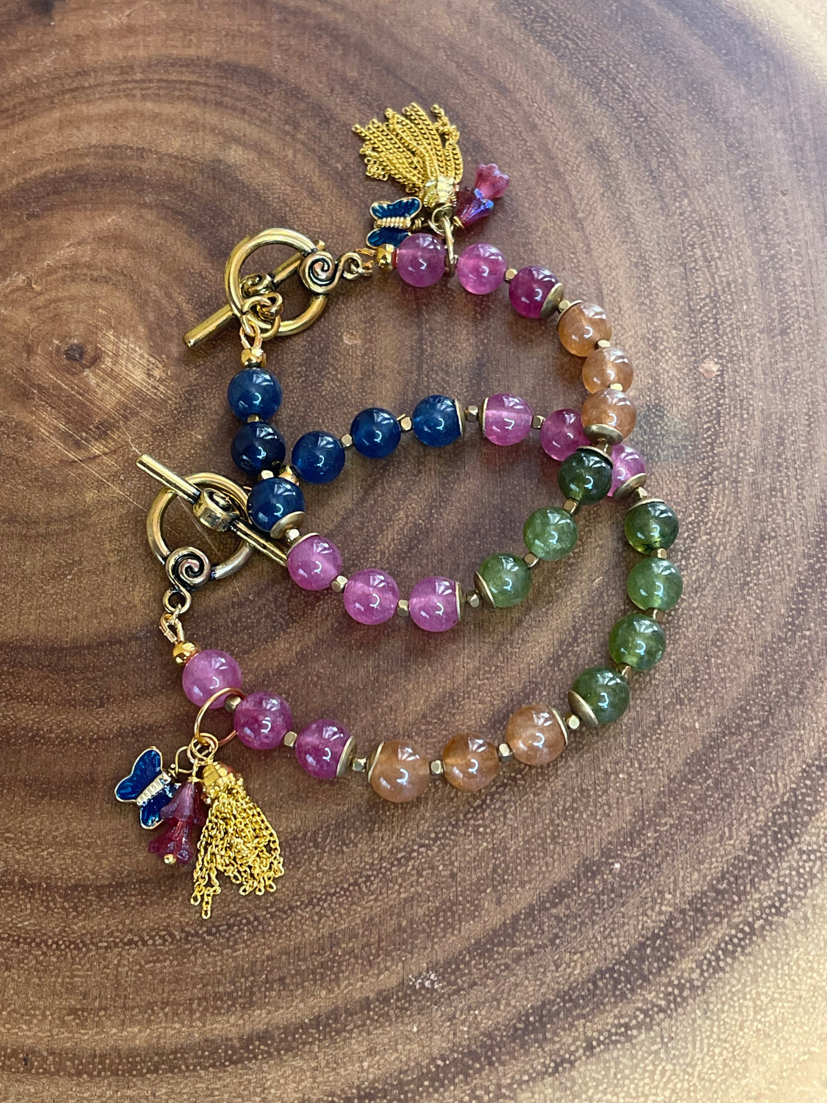 Multicolor natural tourmaline beaded bracelet with an enamel butterfly charm, gold chain tassel, and Czech flower accents, displayed on a wooden surface.