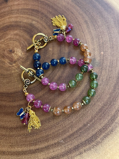 Multicolor natural tourmaline beaded bracelet with an enamel butterfly charm, gold chain tassel, and Czech flower accents, displayed on a wooden surface.