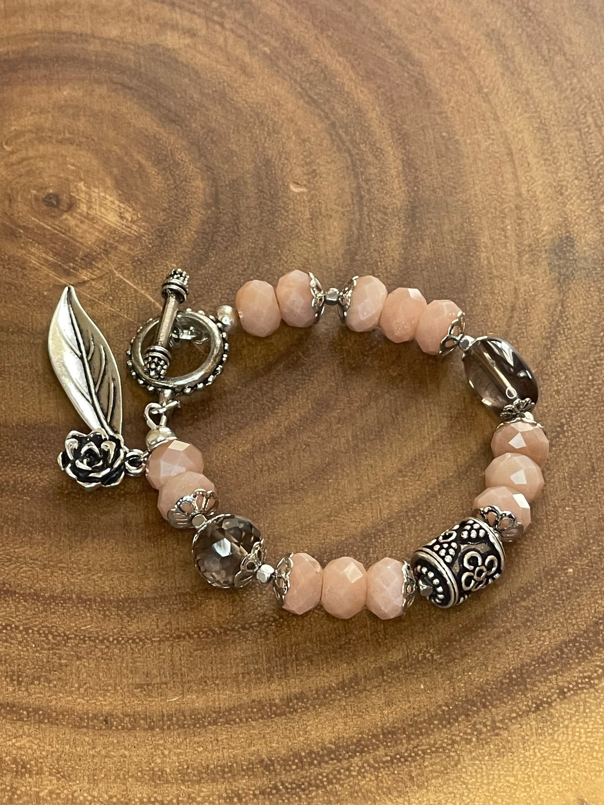 Peach moonstone and smoky quartz beaded bracelet with an ornate silver-plated focal bead, flower and leaf charms, displayed on a wooden surface.