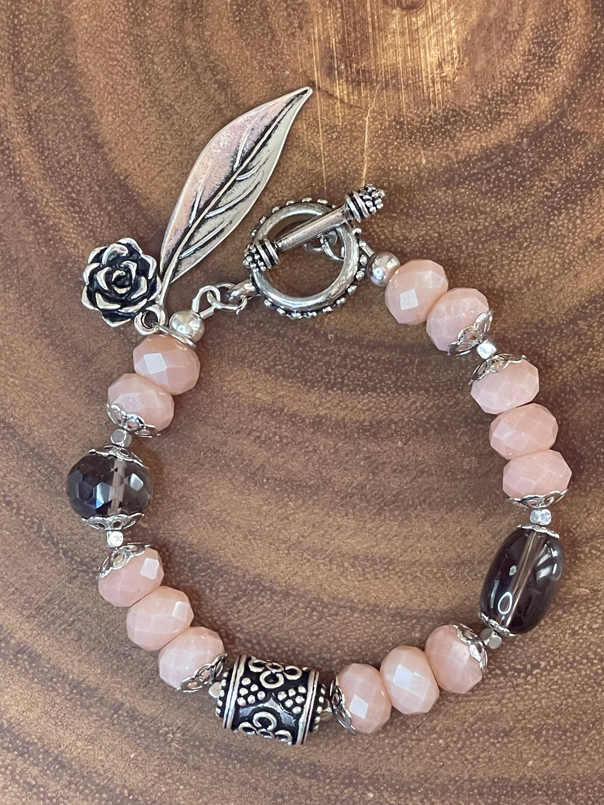 Peach moonstone and smoky quartz beaded bracelet with an ornate silver-plated focal bead, flower and leaf charms, displayed on a wooden surface.