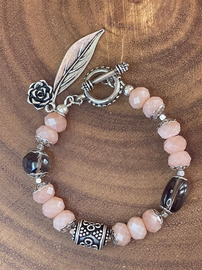 Peach moonstone and smoky quartz beaded bracelet with an ornate silver-plated focal bead, flower and leaf charms, displayed on a wooden surface.