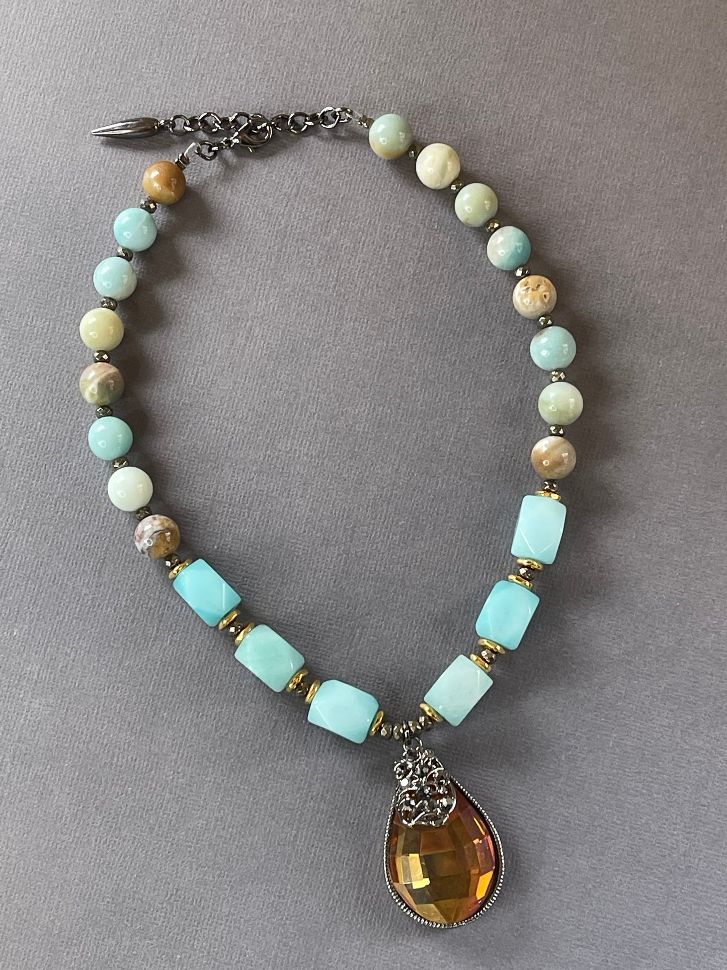 A beaded necklace with light blue, cream, and brown stone beads, accented with gold spacer rings and a large faceted amber teardrop pendant on a gray background.