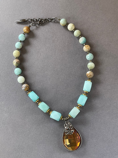 A beaded necklace with light blue, cream, and brown stone beads, accented with gold spacer rings and a large faceted amber teardrop pendant on a gray background.