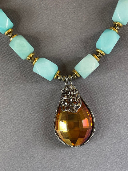 A close-up of a beaded necklace featuring light aqua blue rectangular beads separated by gold-toned spacers, with a large faceted amber-brown teardrop pendant in a silver-toned setting on a gray textured background.