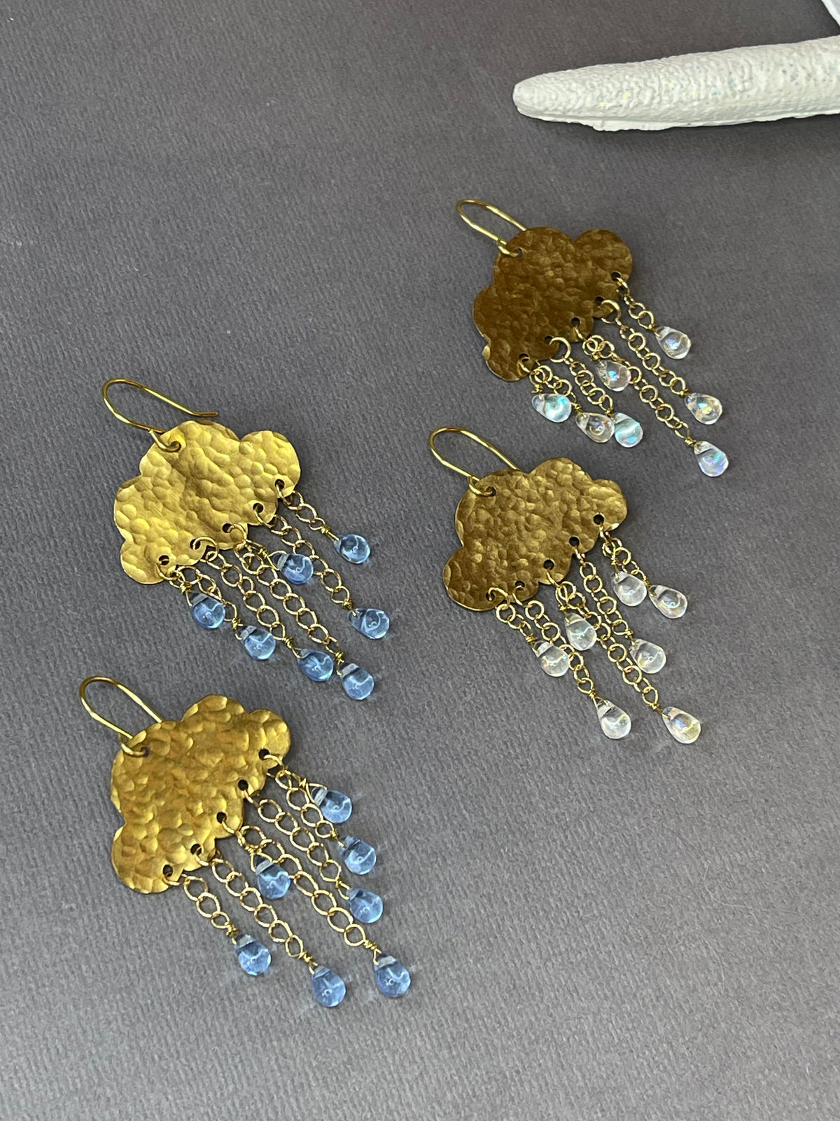 Gold-tone rain cloud dangle earrings with textured brass clouds and dangling raindrop accents on a gray background.
