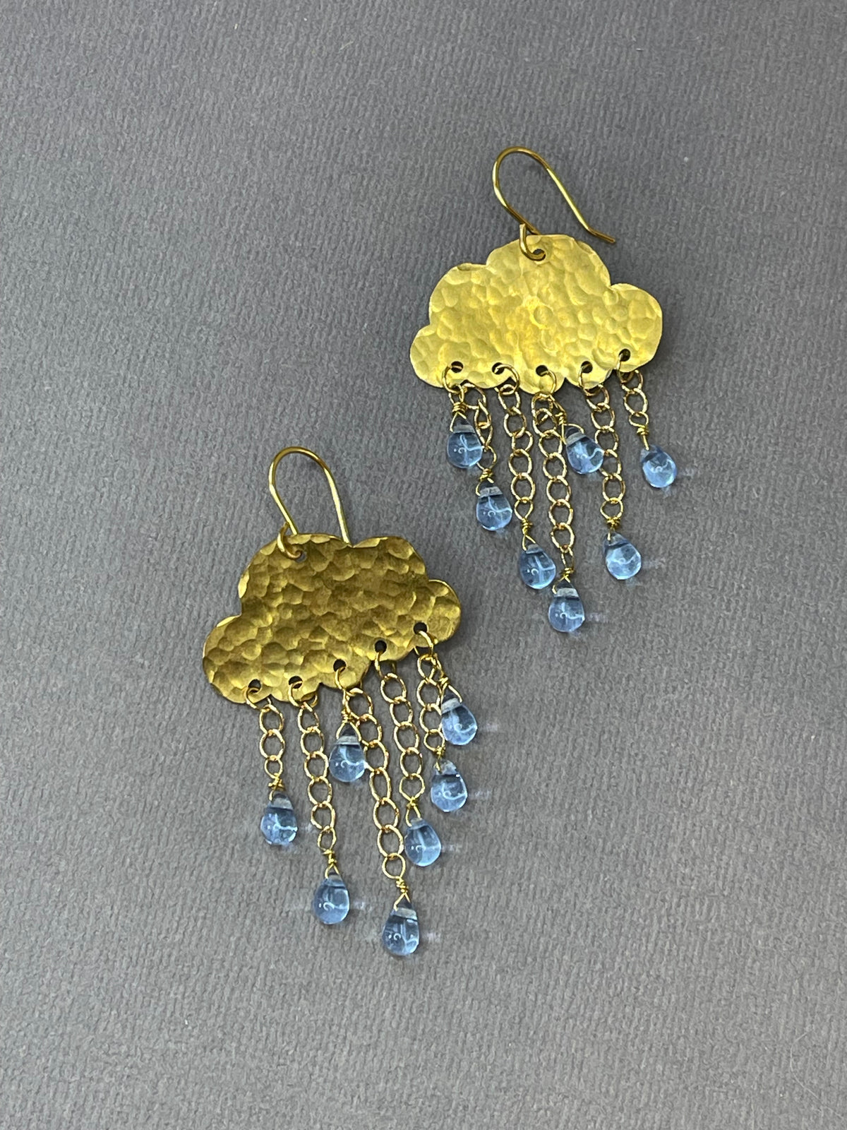 Gold-tone rain cloud dangle earrings with textured brass clouds and dangling raindrop accents on a gray background.