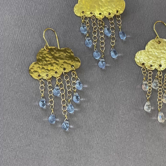 Gold-tone rain cloud dangle earrings with textured brass clouds and dangling raindrop accents on a gray background.