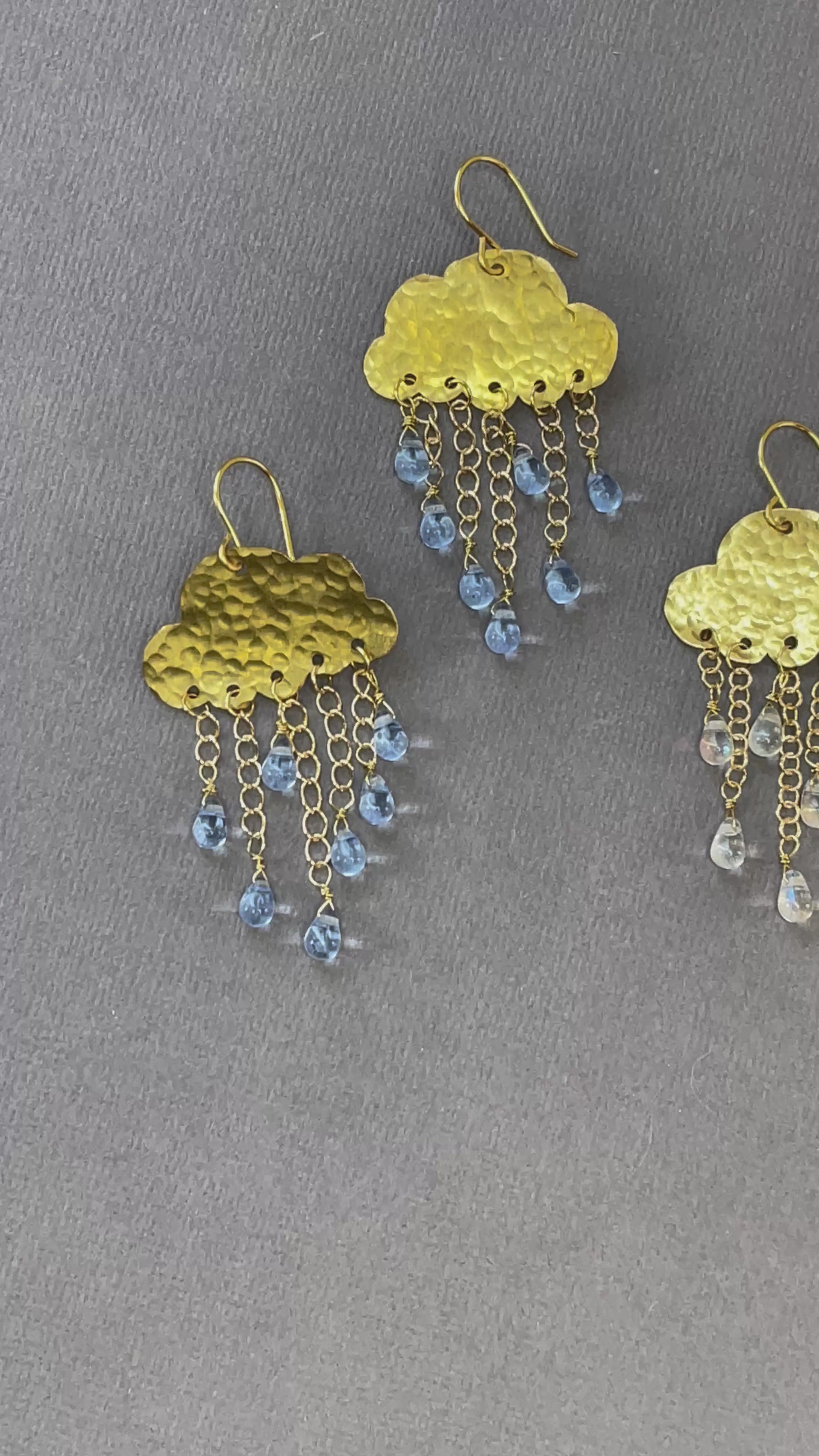 Gold-tone rain cloud dangle earrings with textured brass clouds and dangling raindrop accents on a gray background.