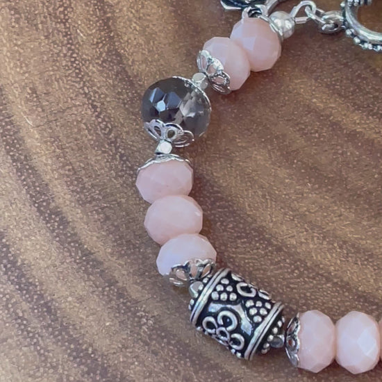 Peach moonstone and smoky quartz beaded bracelet with an ornate silver-plated focal bead, flower and leaf charms, displayed on a wooden surface.