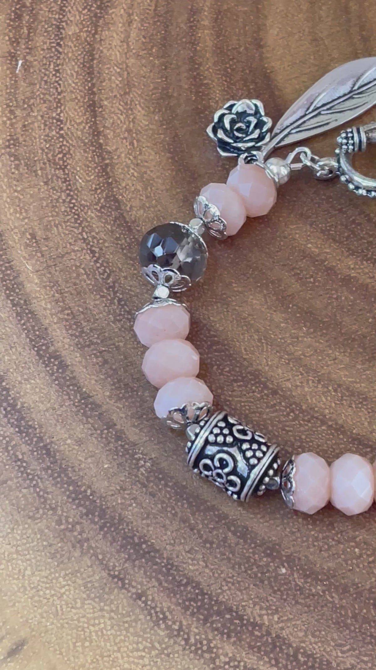 Peach moonstone and smoky quartz beaded bracelet with an ornate silver-plated focal bead, flower and leaf charms, displayed on a wooden surface.