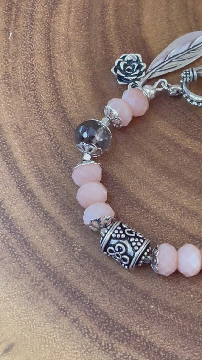Peach moonstone and smoky quartz beaded bracelet with an ornate silver-plated focal bead, flower and leaf charms, displayed on a wooden surface.