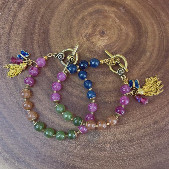 Multicolor natural tourmaline beaded bracelet with an enamel butterfly charm, gold chain tassel, and Czech flower accents, displayed on a wooden surface.