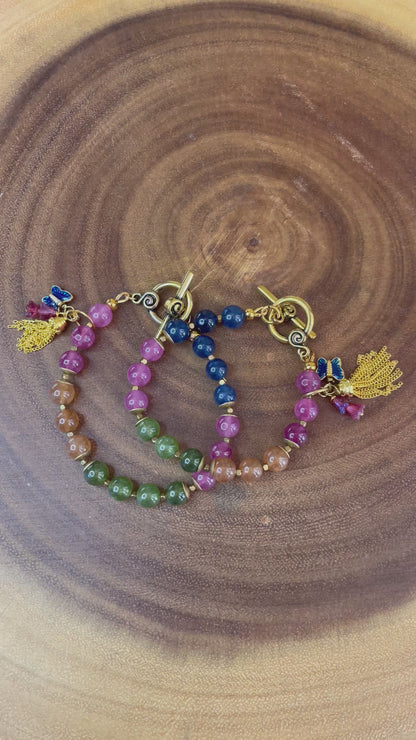 Multicolor natural tourmaline beaded bracelet with an enamel butterfly charm, gold chain tassel, and Czech flower accents, displayed on a wooden surface.